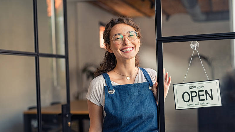 Smiling woman at the entrance to her business with an open sign.